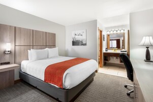A hotel room with a bed, bedside table, lamp, desk, chair, and a bathroom with a mirror and sink.