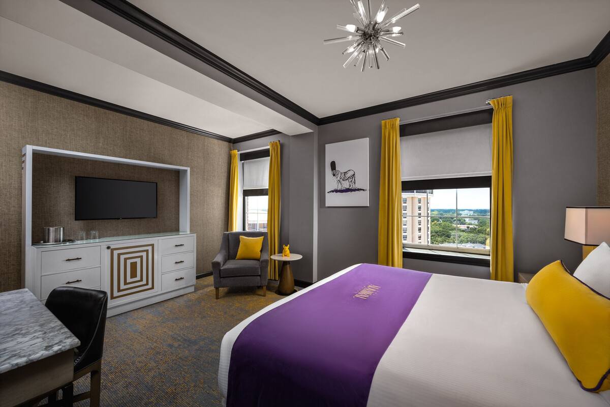 Habitación de hotel con cama, televisión montada en un mueble, escritorio, silla y ventana con cortinas.