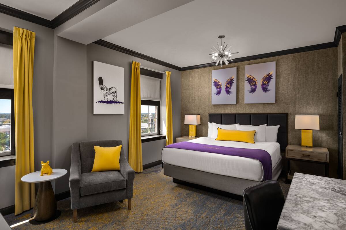 Una habitación de hotel con una cama, un sofá gris con un cojín amarillo, una mesita con una figurita amarilla y obras de arte en las paredes.