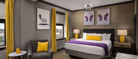 A hotel room with a bed, a gray sofa with a yellow pillow, a small table with a yellow figurine, and artwork on the walls.