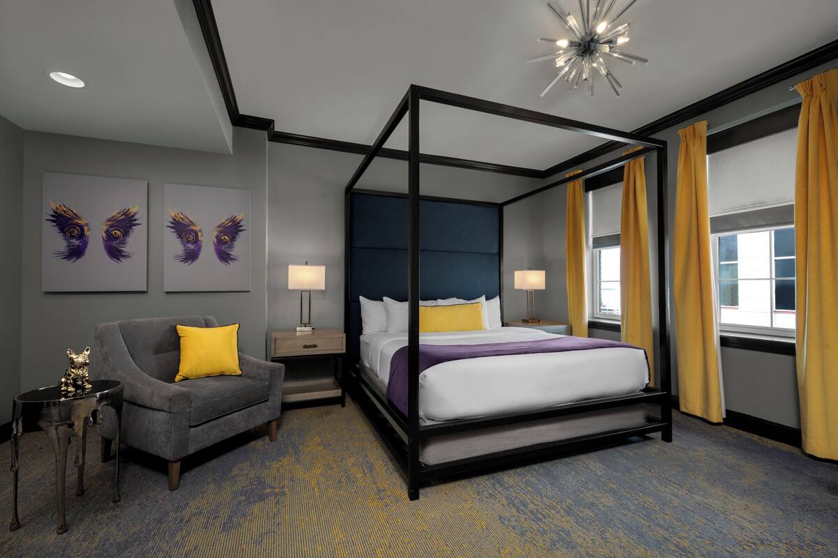 Una habitación de hotel moderna con una cama con dosel, un sofá gris con un cojín amarillo y obras de arte en la pared.