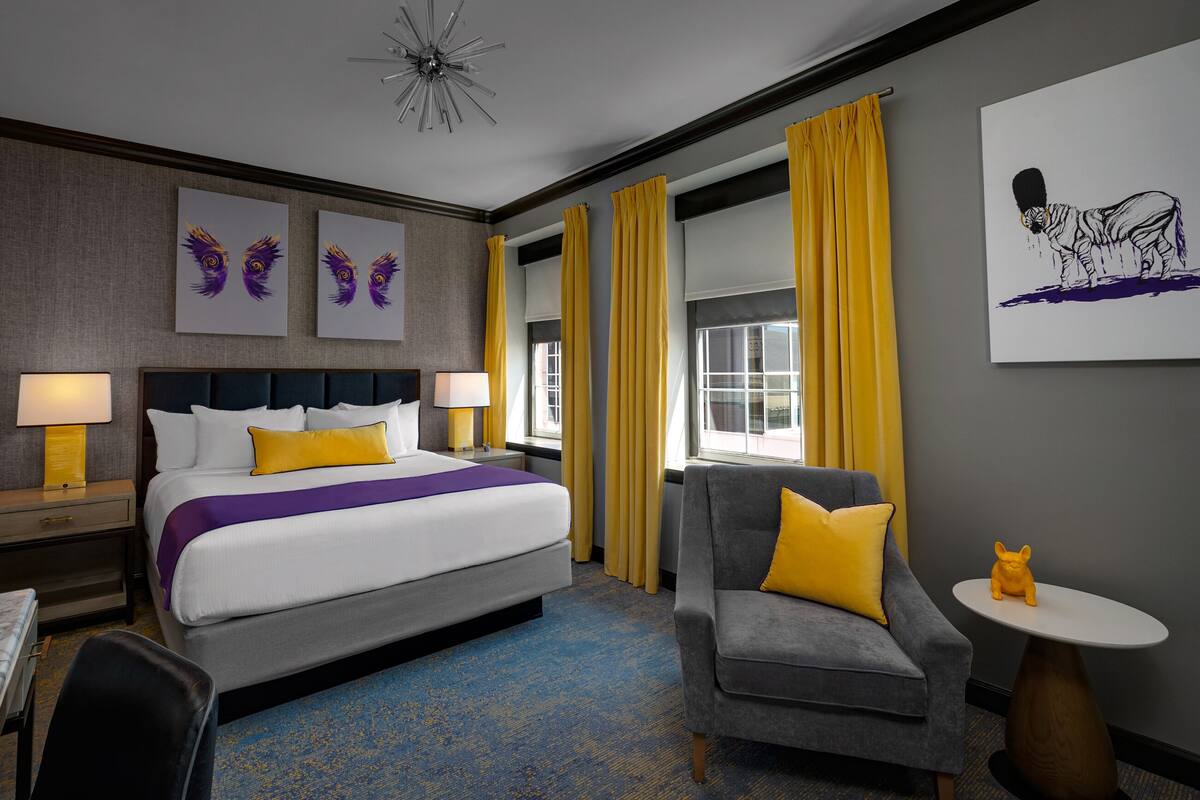 Un cuarto de hotel con una cama grande, dos mesitas de noche con lámparas, un sillón gris, una pequeña mesa redonda con una figurita y obras de arte en las paredes.
