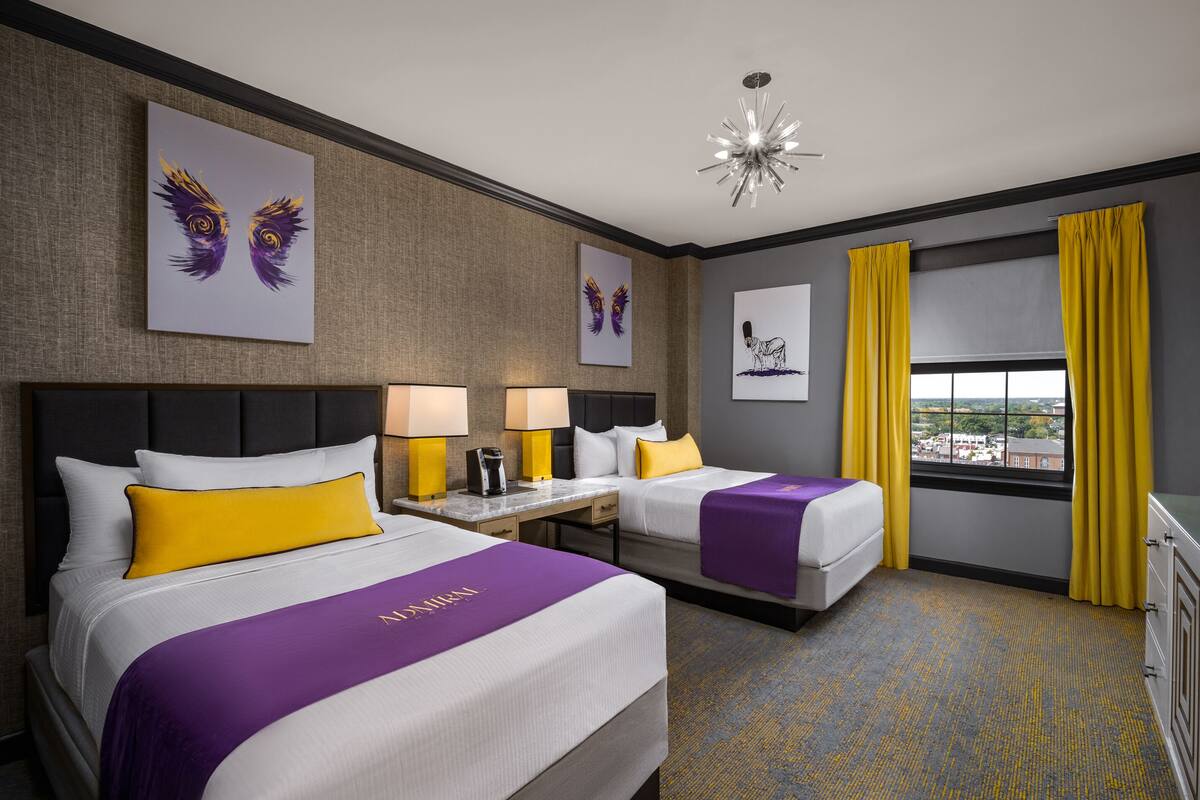 Habitación de hotel con dos camas, un escritorio y una ventana con cortinas amarillas.