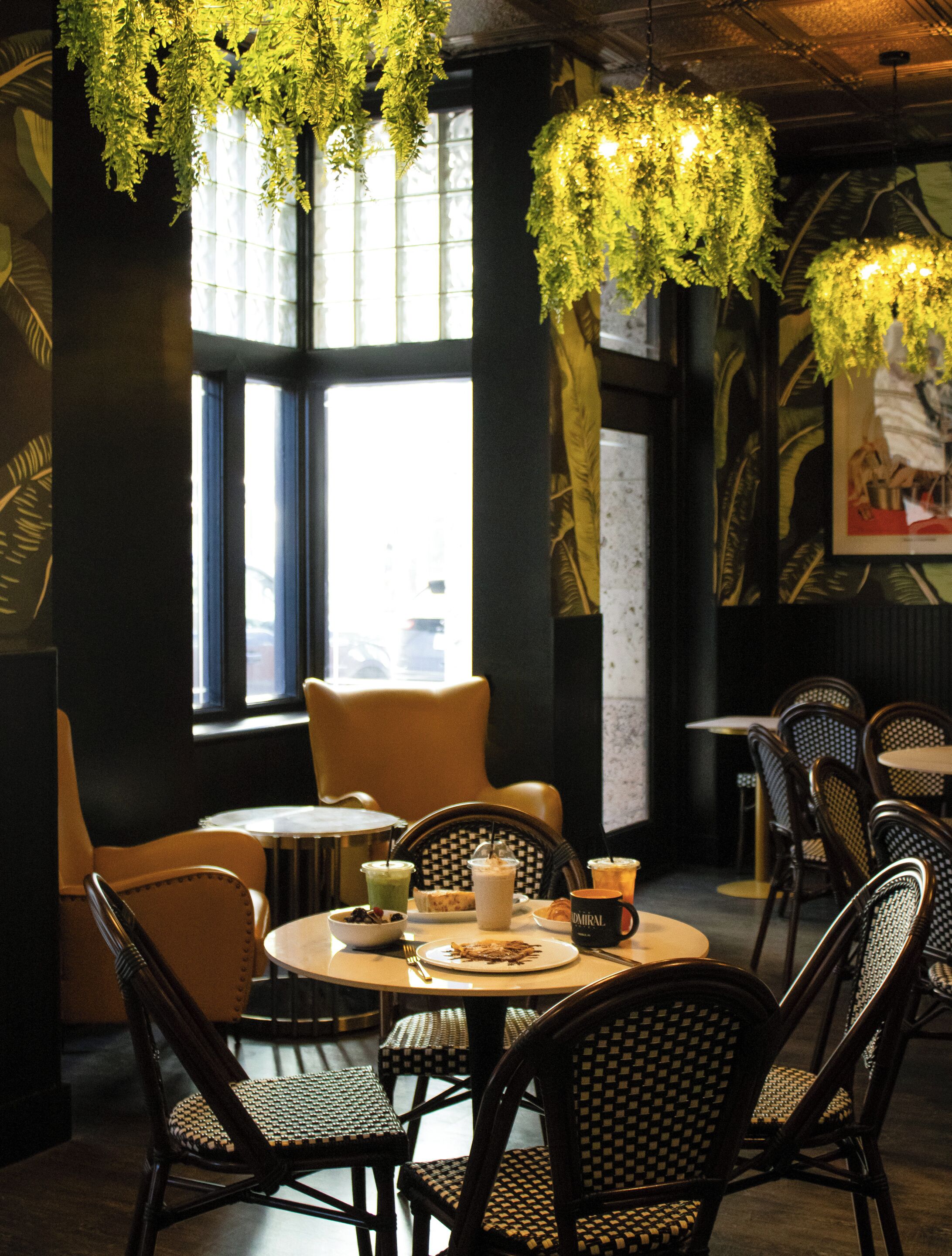 A restaurant interior with round tables, wicker chairs, hanging plants, and a view of the city through large windows.