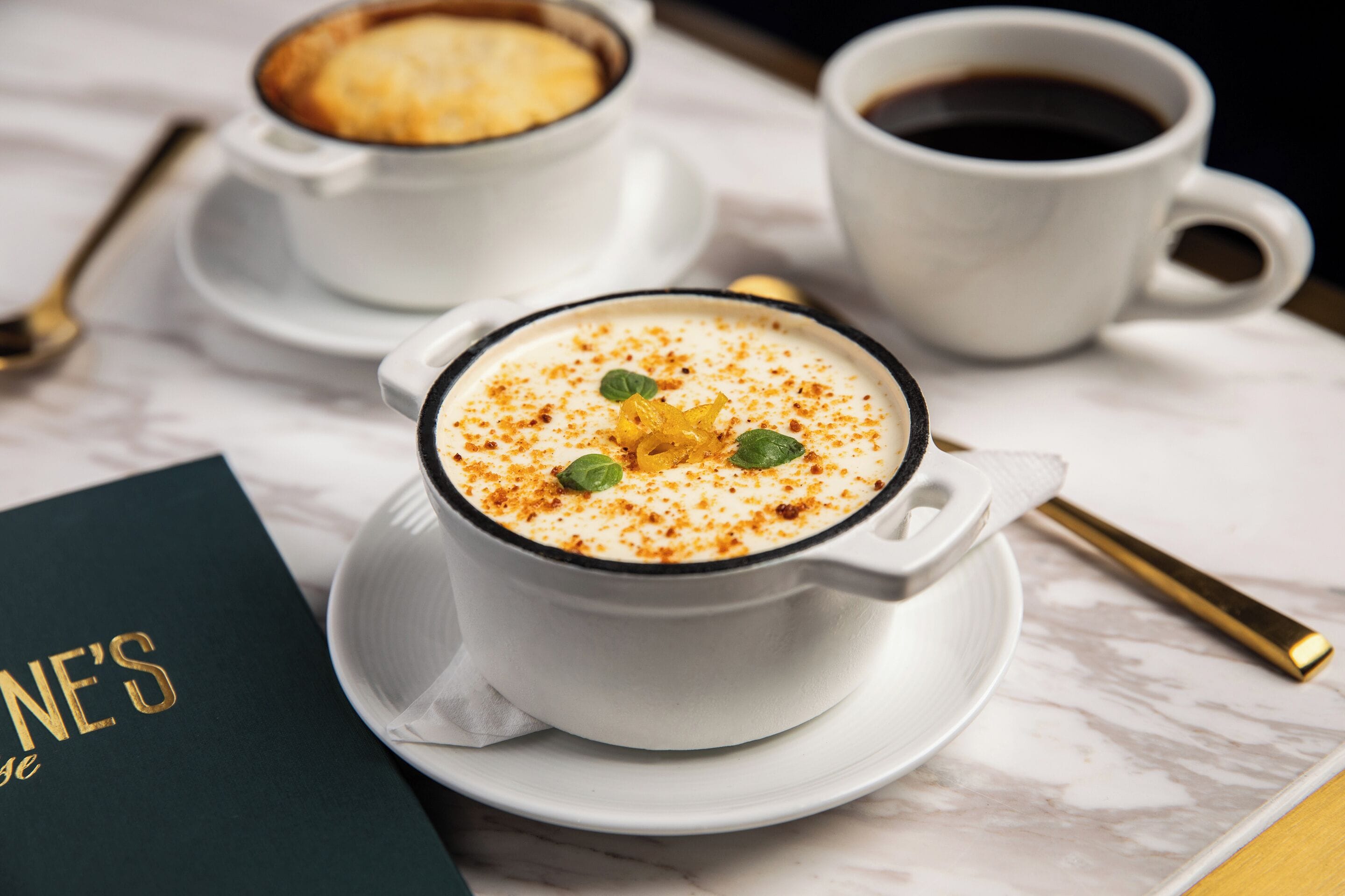 Un postre en una taza blanca con plato, una taza de café y un menú sobre una mesa de mármol.