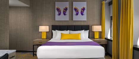 A hotel room with a large bed, two wall-mounted artworks, a nightstand with a lamp, and a window with yellow curtains.