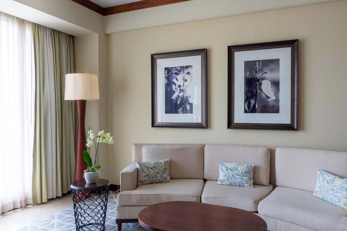A living room with a beige sofa, two framed black and white floral paintings, a round wooden coffee table, a side table with a lamp, and a potted orchid.