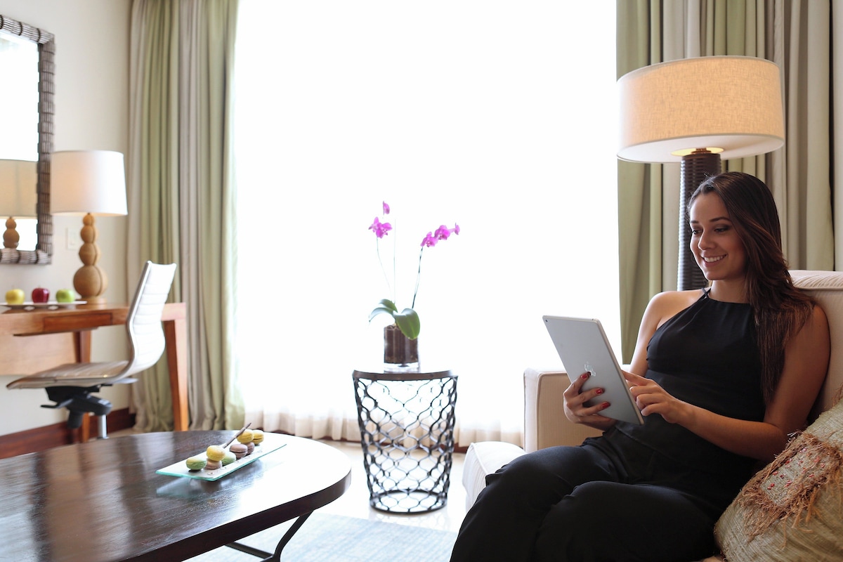 A woman sitting on a couch, holding a tablet. There is a table with a plate of food and a vase with flowers. A lamp and a mirror are on the wall.