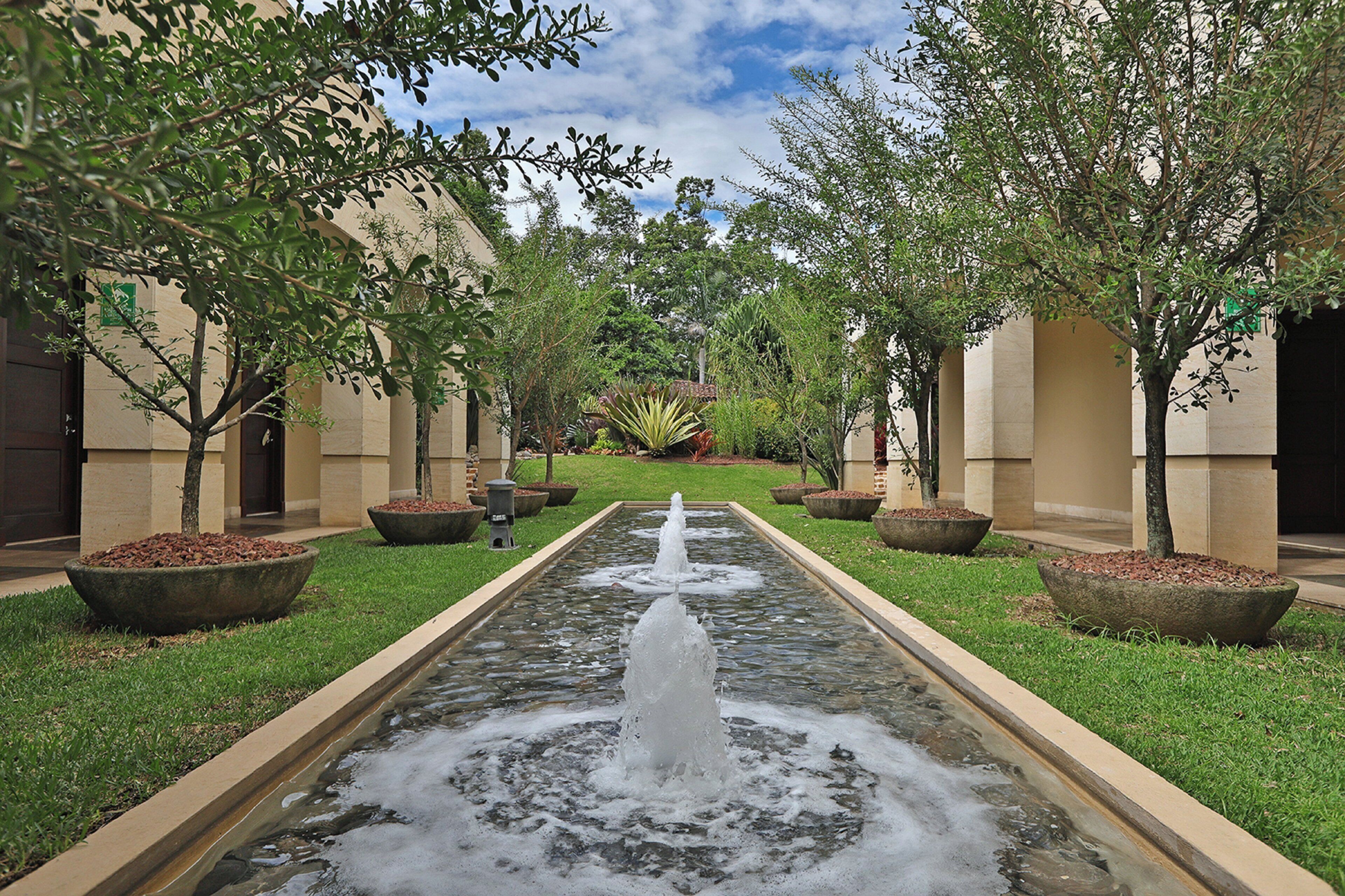 A courtyard with a central water feature, surrounded by trees and planters, and buildings with beige walls and columns.