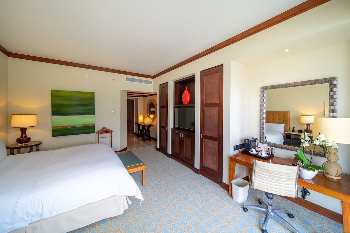 A hotel room with a large bed, a desk with a computer, a mirror, and a potted plant.