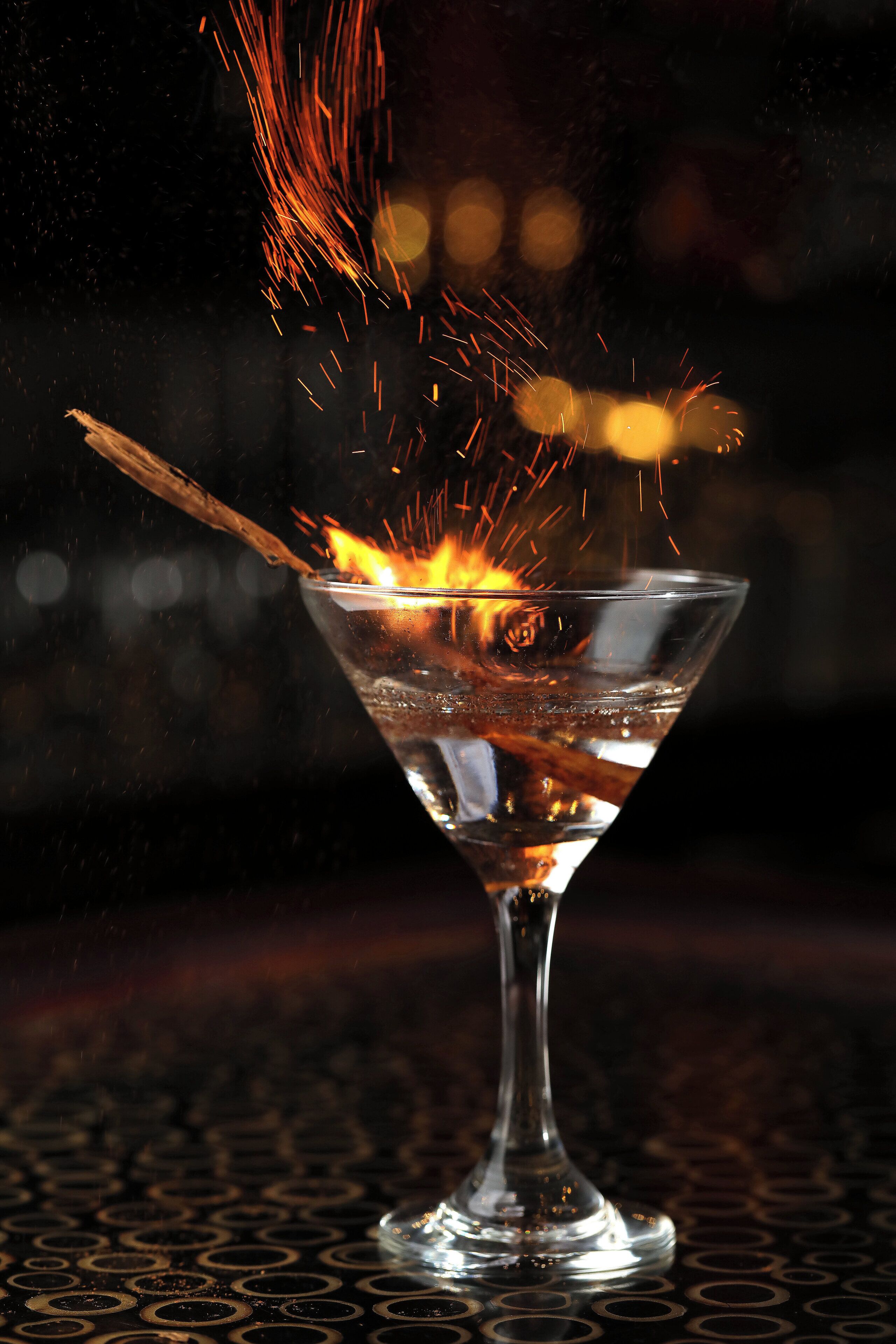Una copa de martini con una pajita de madera y un chorro de líquido, posiblemente un cóctel, con chispas que suben.