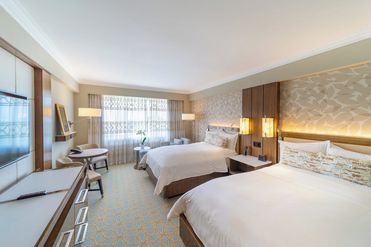 A hotel room with two beds, a desk, a chair, a small table, and a TV.