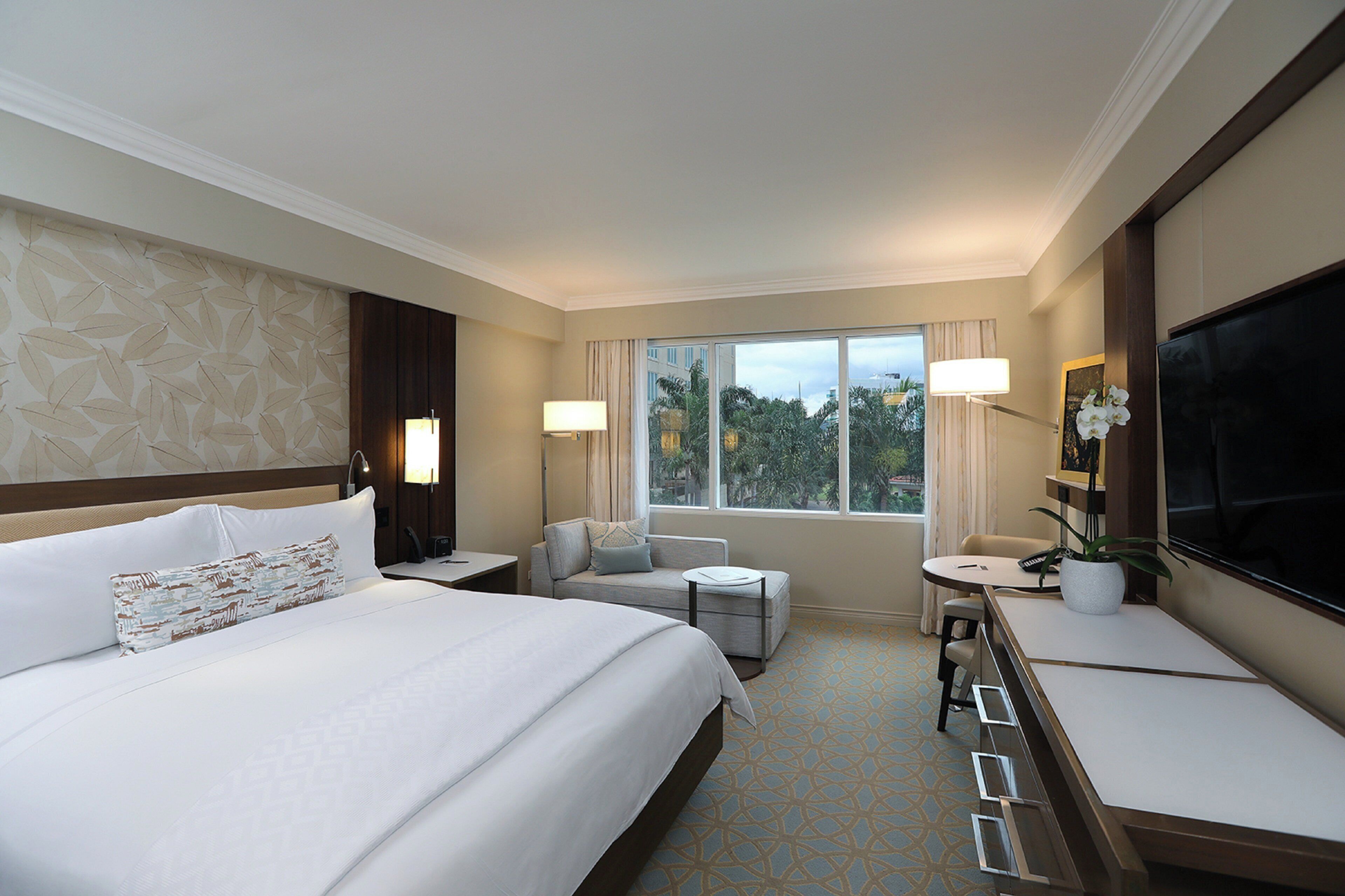 A hotel room with a large bed, a desk, a chair, a TV, and a window with a view of trees.