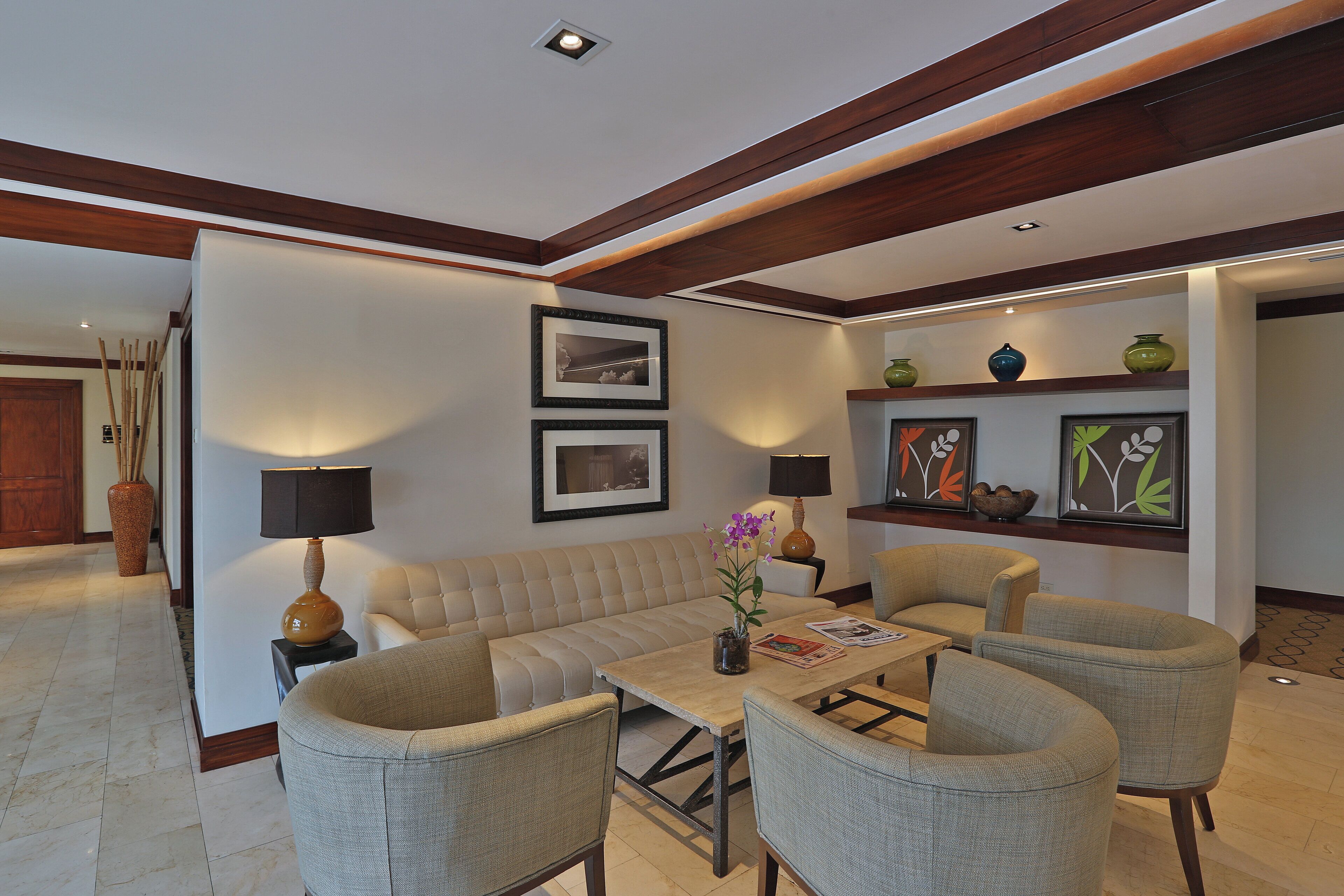 A modern living room with a beige sofa, two armchairs, a coffee table, and a fireplace. There are framed pictures on the wall, a vase with flowers, and a lamp on a side table.