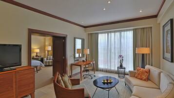 A hotel room with a television, a sofa, a coffee table, a desk, and a chair. There is a window with curtains and a lamp on the desk.