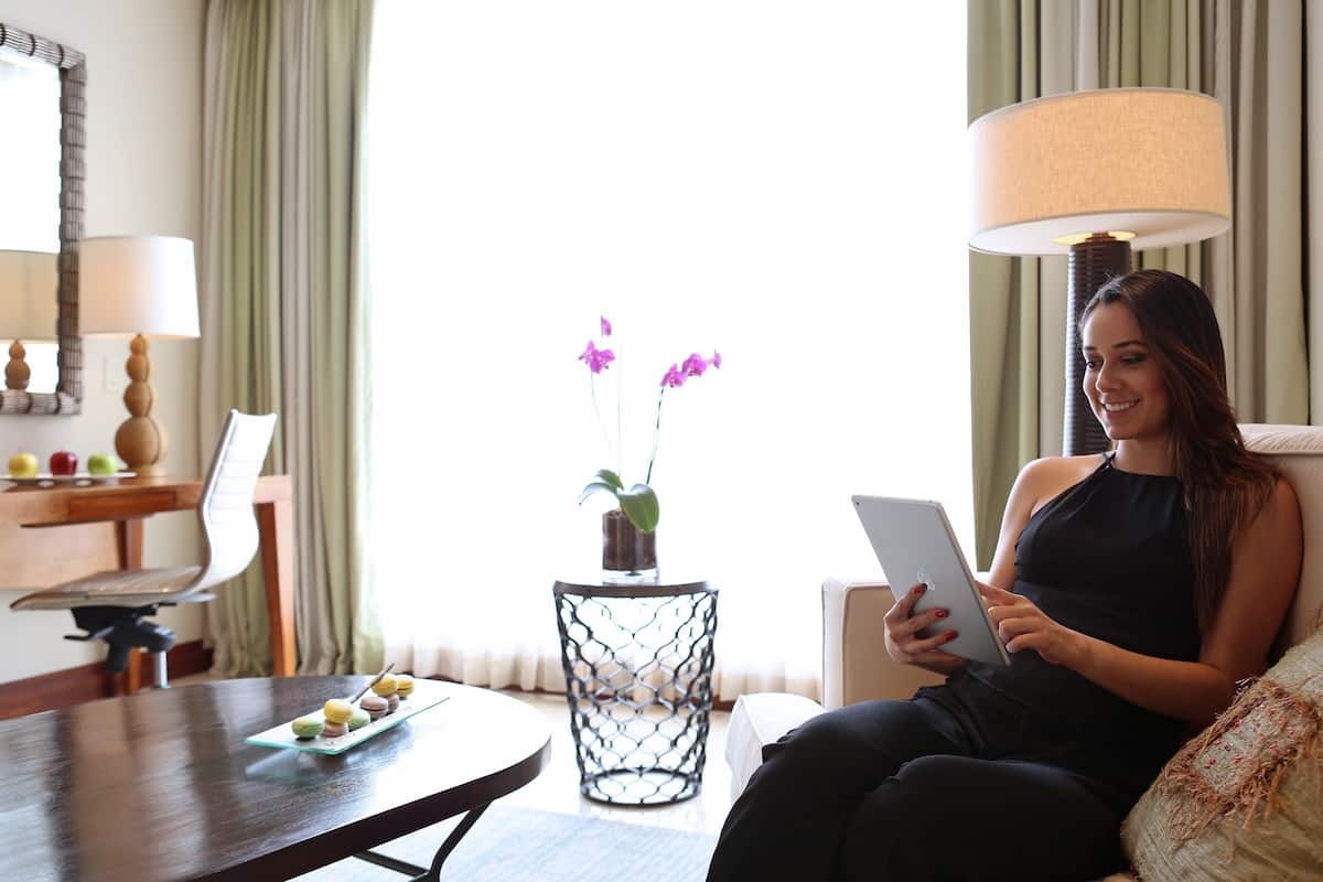 A woman sitting on a couch, holding a tablet. There is a table with a plate of food and a vase with flowers. A lamp and a mirror are on the wall.