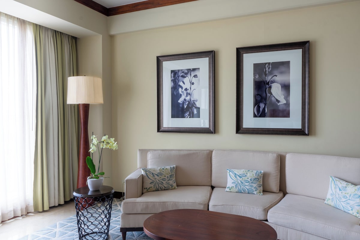 A living room with a beige sofa, two framed black and white floral paintings, a round wooden coffee table, a side table with a lamp, and a potted orchid.
