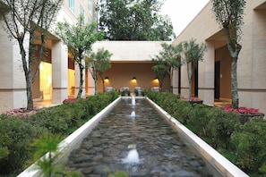 Un élément décoratif aquatique, agrémenté d’un sentier bordé de pierres, flanqué d’arbres et d’arbustes, et menant à l’entrée d’un bâtiment.