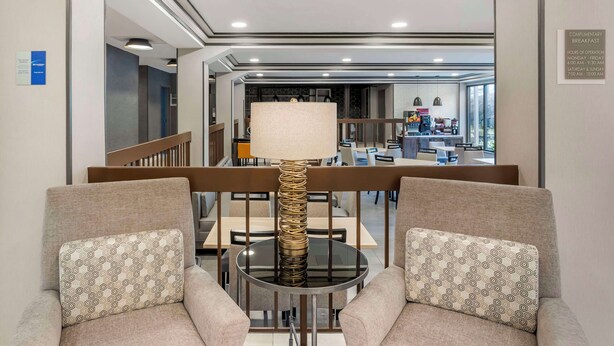 A modern hotel lobby with a seating area featuring two sofas with patterned cushions, a central round table with a unique lamp, and a view into the dining area.