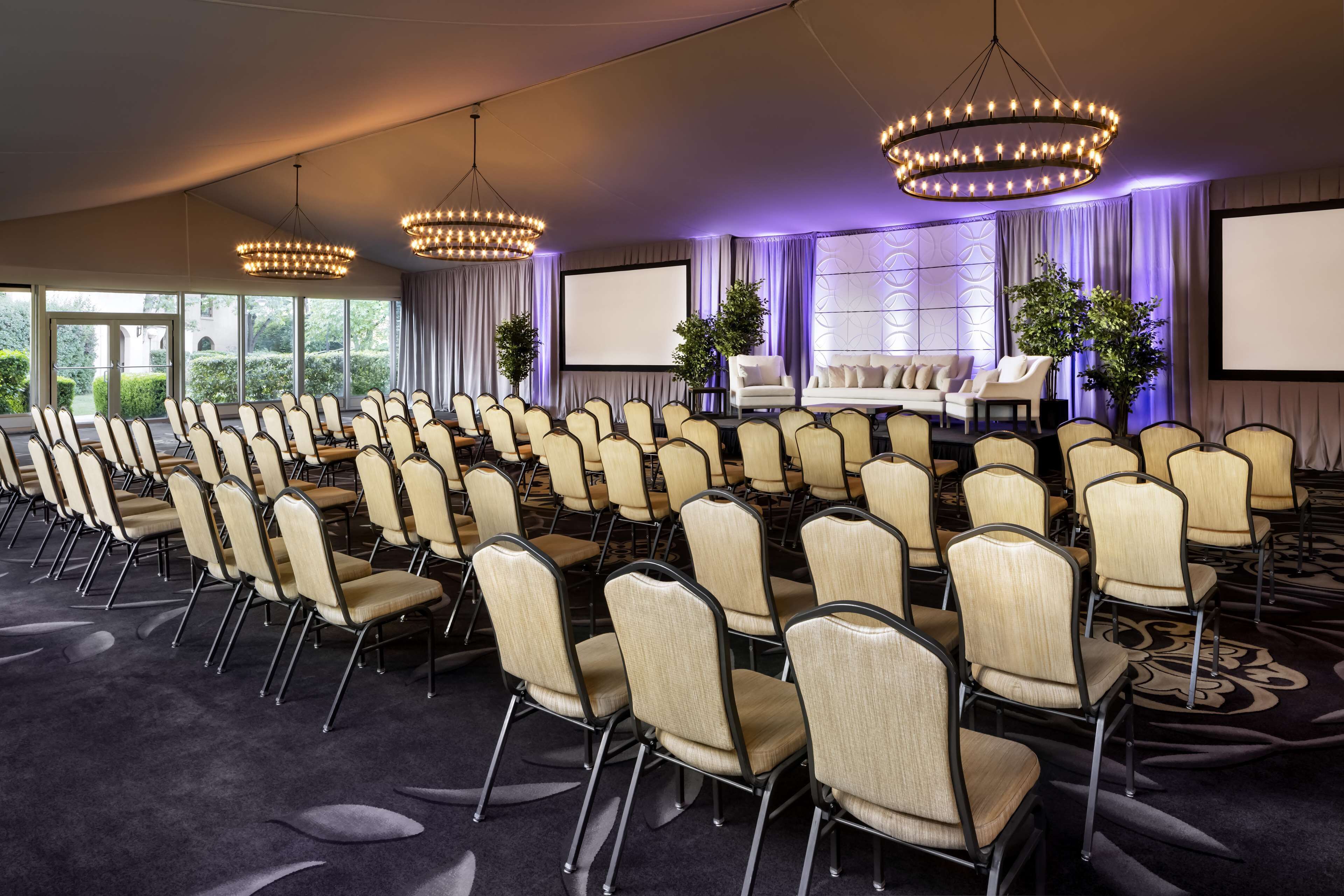 Un salón de conferencias con filas de sillas, un escenario con una pantalla y ventanas amplias.
