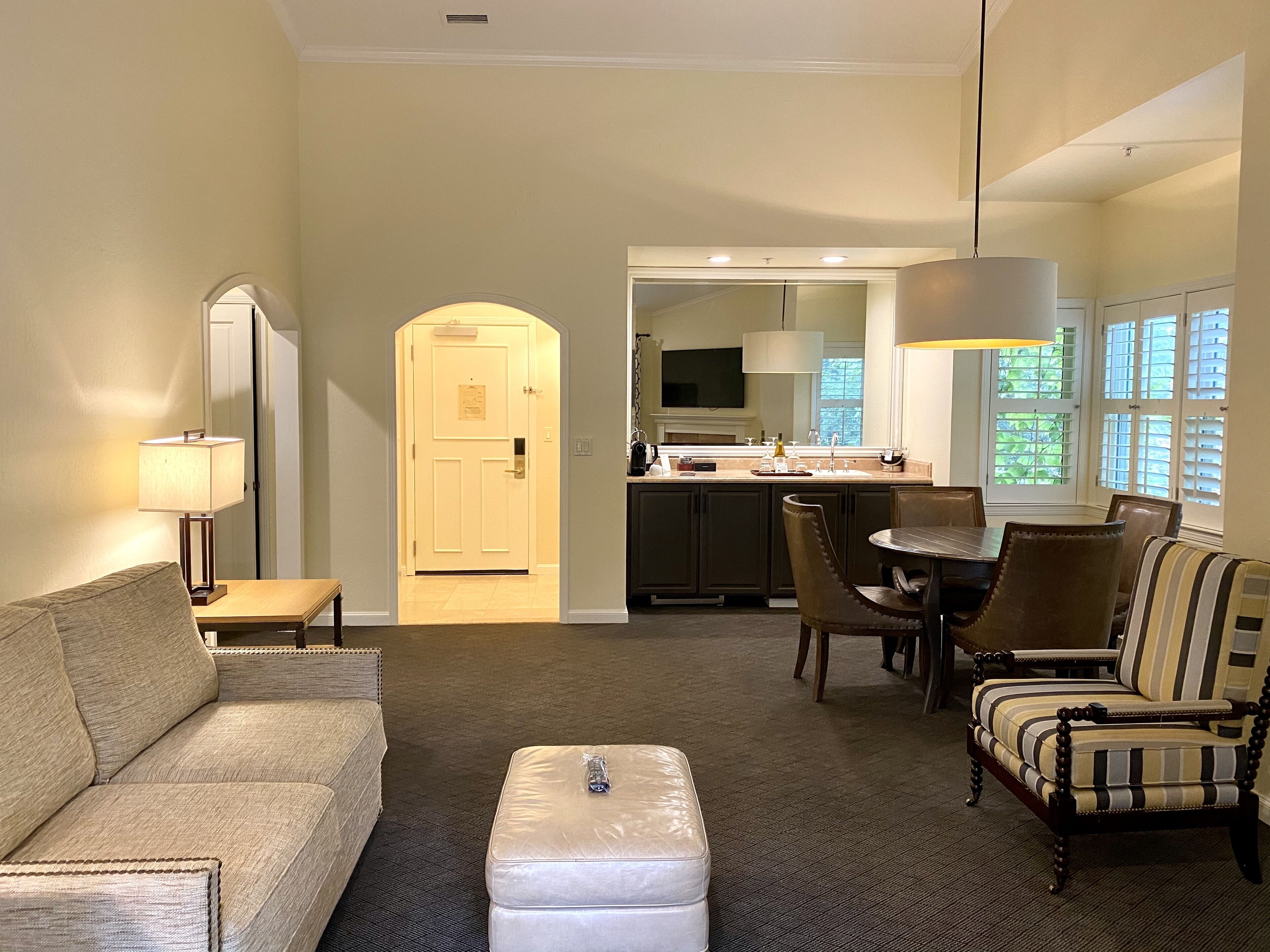 A living room with a sofa, ottoman, and striped armchairs, a dining area with a table and chairs, a kitchen with a TV, and a hallway leading to a door.