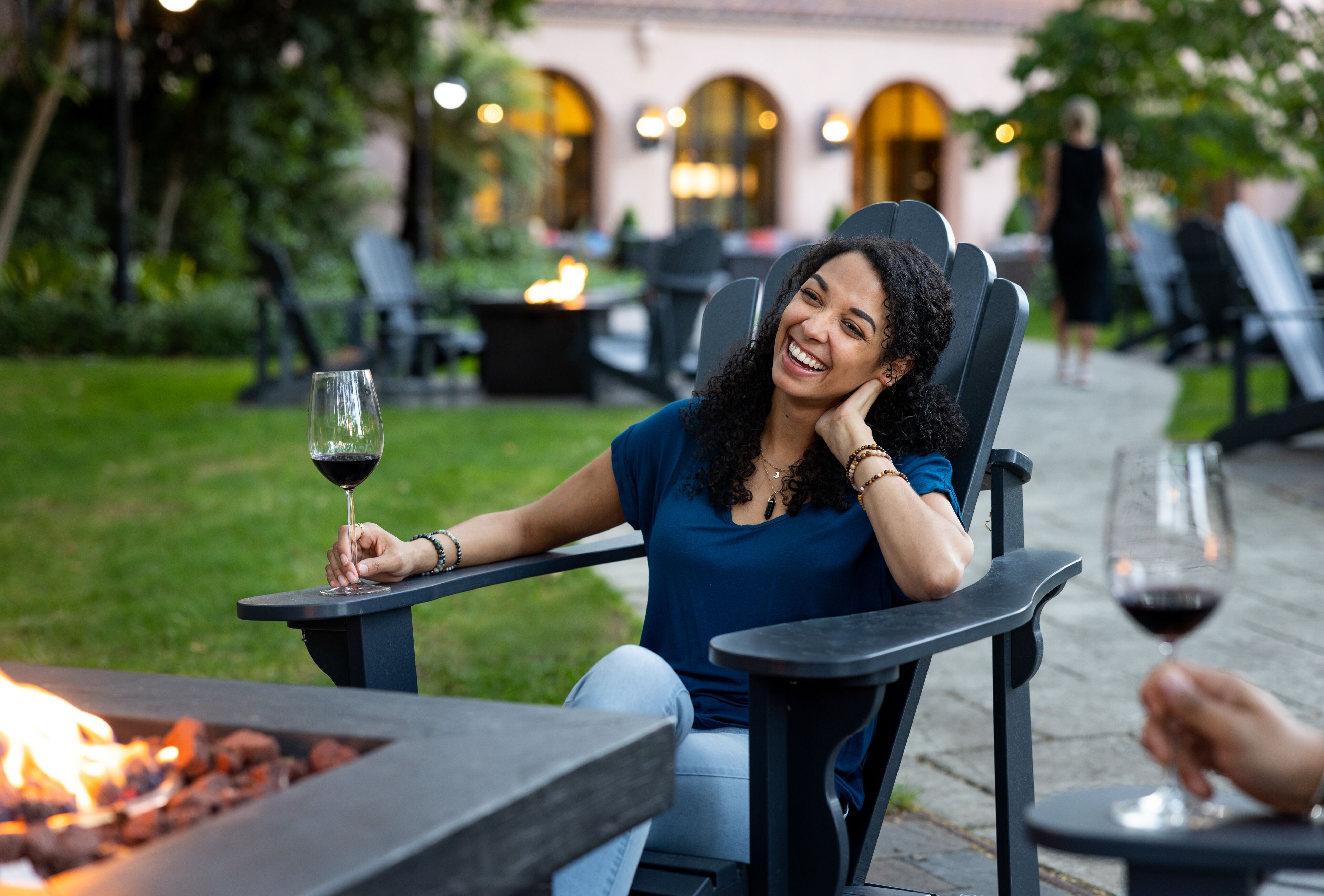A person seated at a table with a glass of wine, a fire feature in the foreground, and outdoor seating in a garden setting.