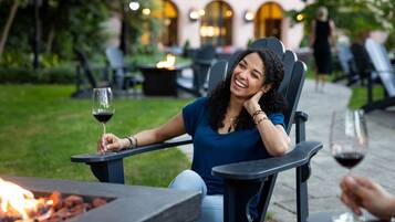 A person seated at a table with a glass of wine, a fire feature in the foreground, and outdoor seating in a garden setting.