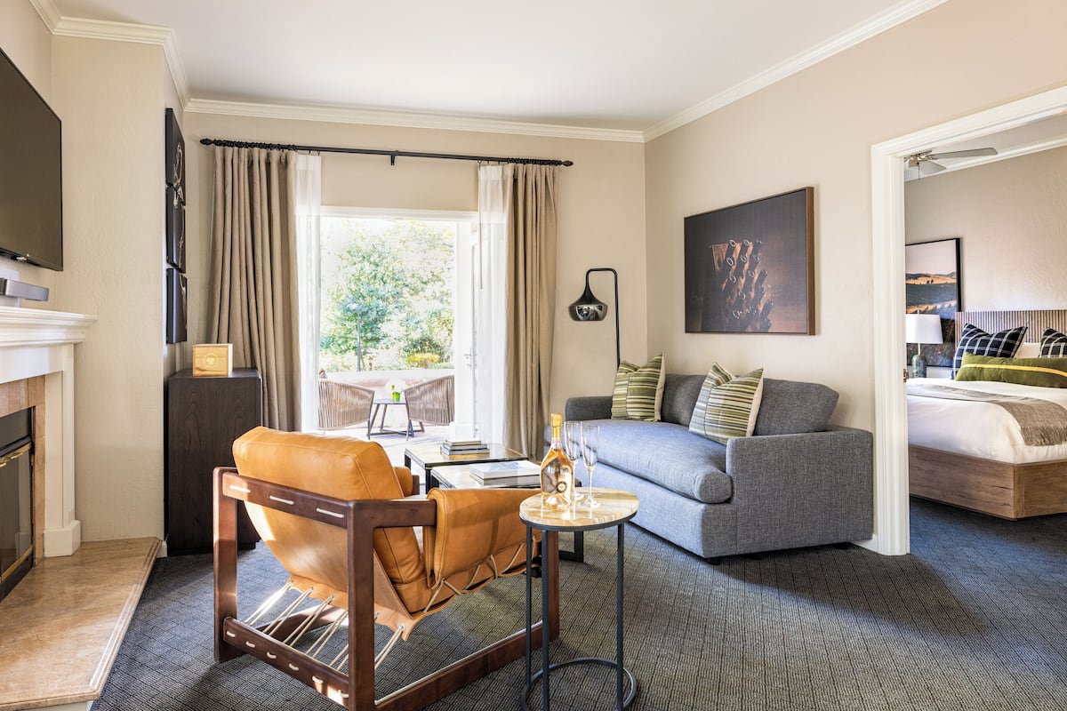 Habitación de hotel con sofá, una silla, una mesita con una botella y vasos, chimenea y una cama.