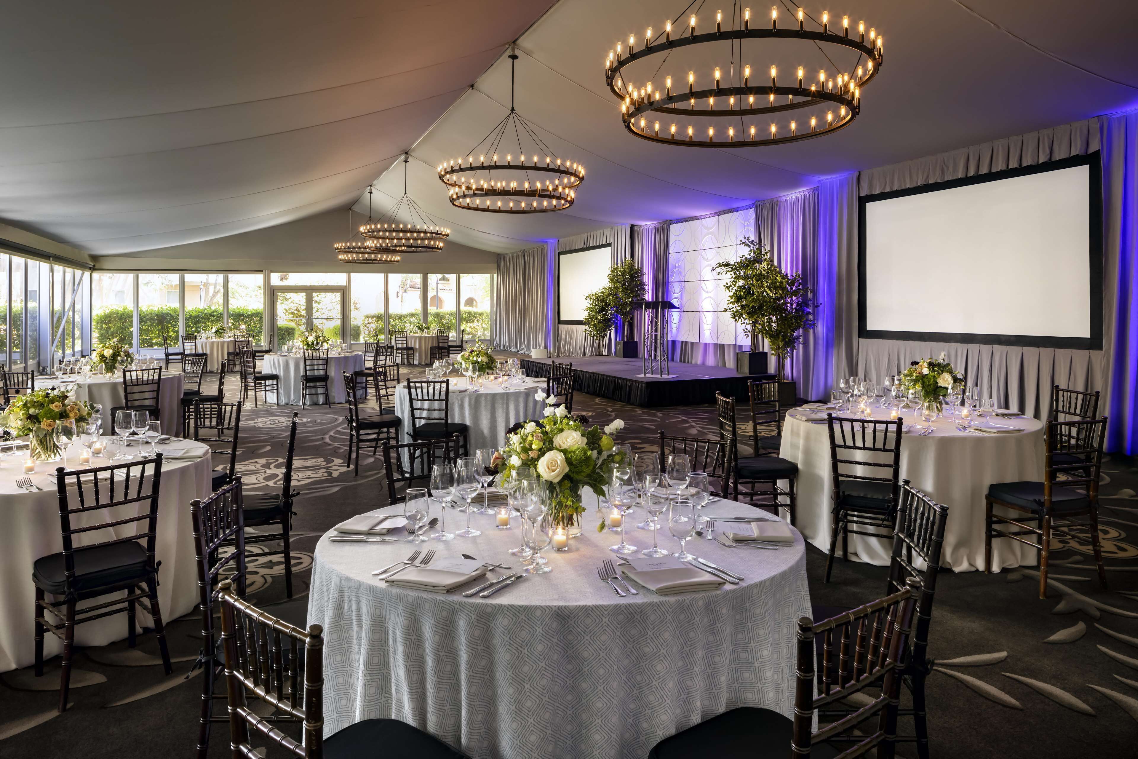 A banquet hall with round tables set for dining, chandeliers, and a stage with curtains.