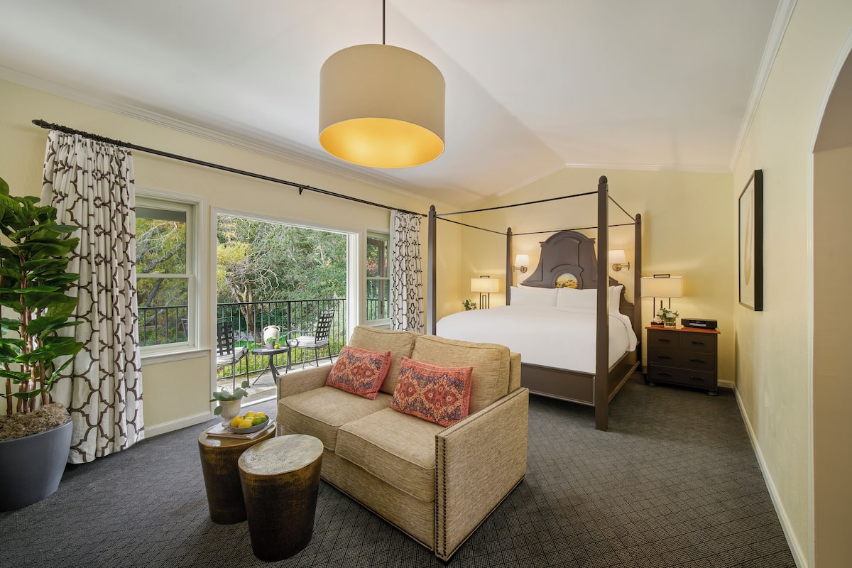 Habitación de hotel con cama con dosel, un sofá con cojines decorativos, una mesita de noche con lámpara y una planta en maceta grande.