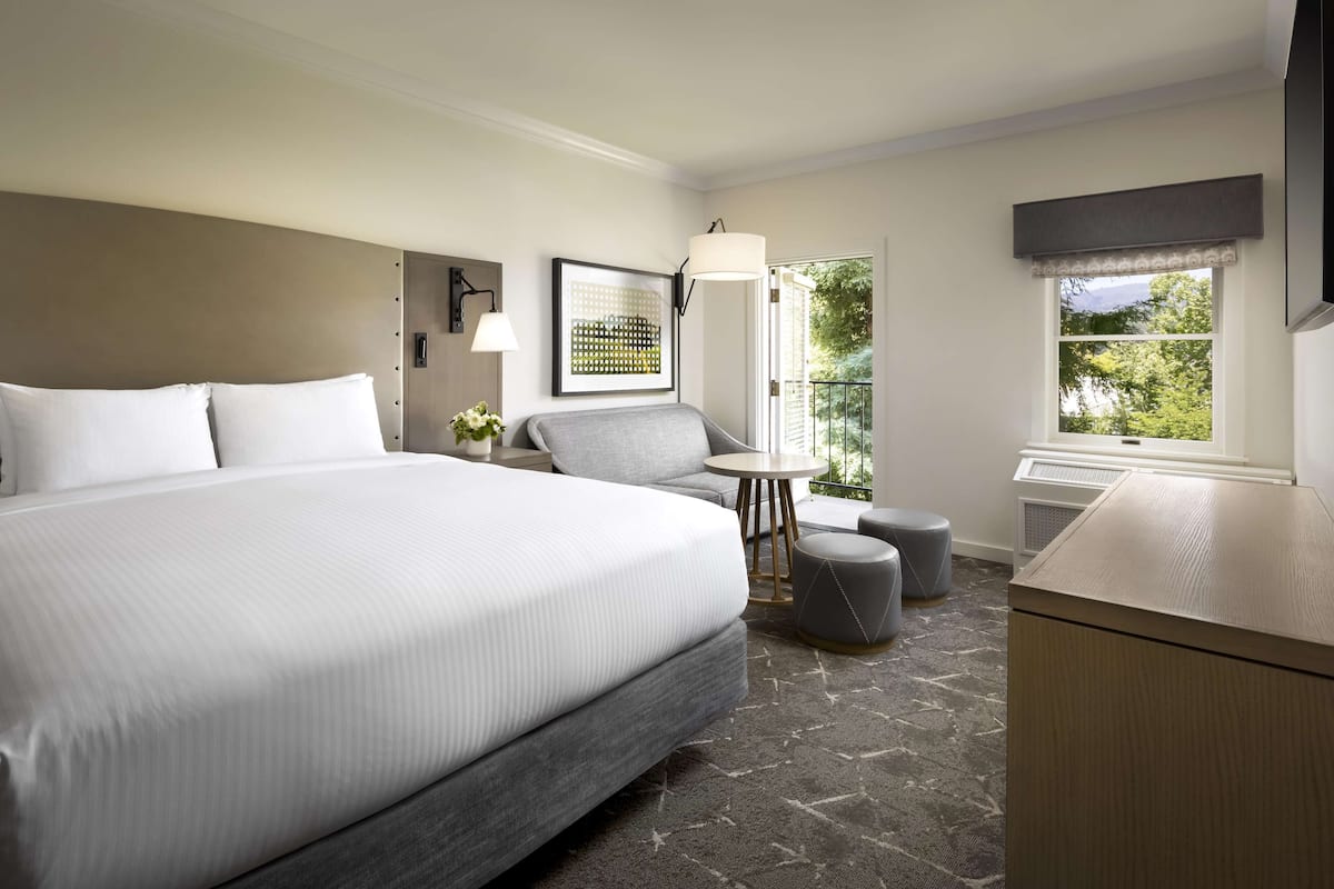 Habitación de hotel con una cama grande, un sillón gris, una mesita redonda y vistas a zonas verdes por la ventana.