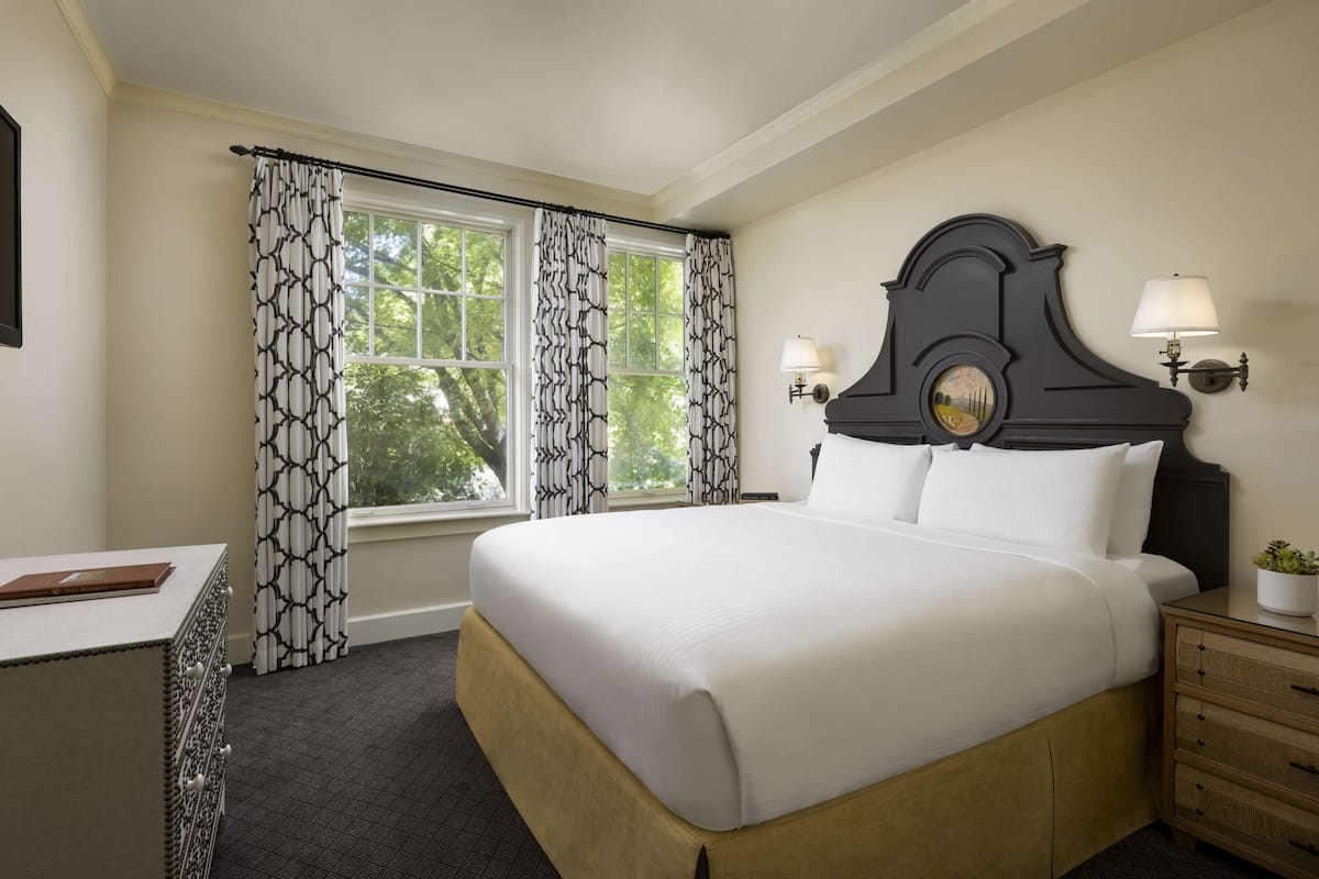 Habitación de hotel ordenada con una cama grande, una mesita de noche, una lámpara y una ventana con cortinas estampadas.