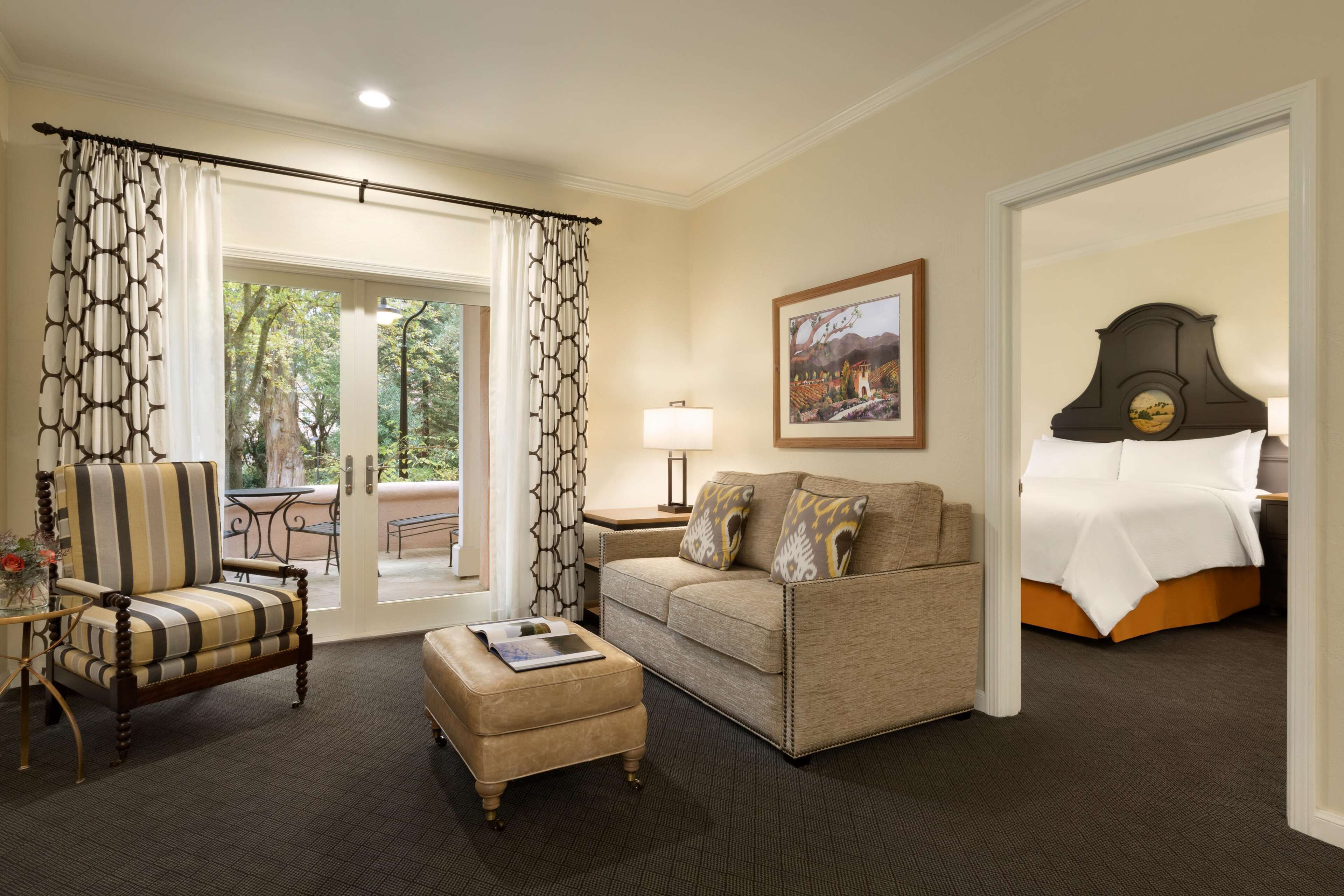 A hotel room with a sofa, armchair, and ottoman, a bed, a nightstand with a lamp, and a painting on the wall.