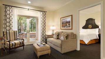 A hotel room with a sofa, armchair, and ottoman, a bed, a nightstand with a lamp, and a painting on the wall.