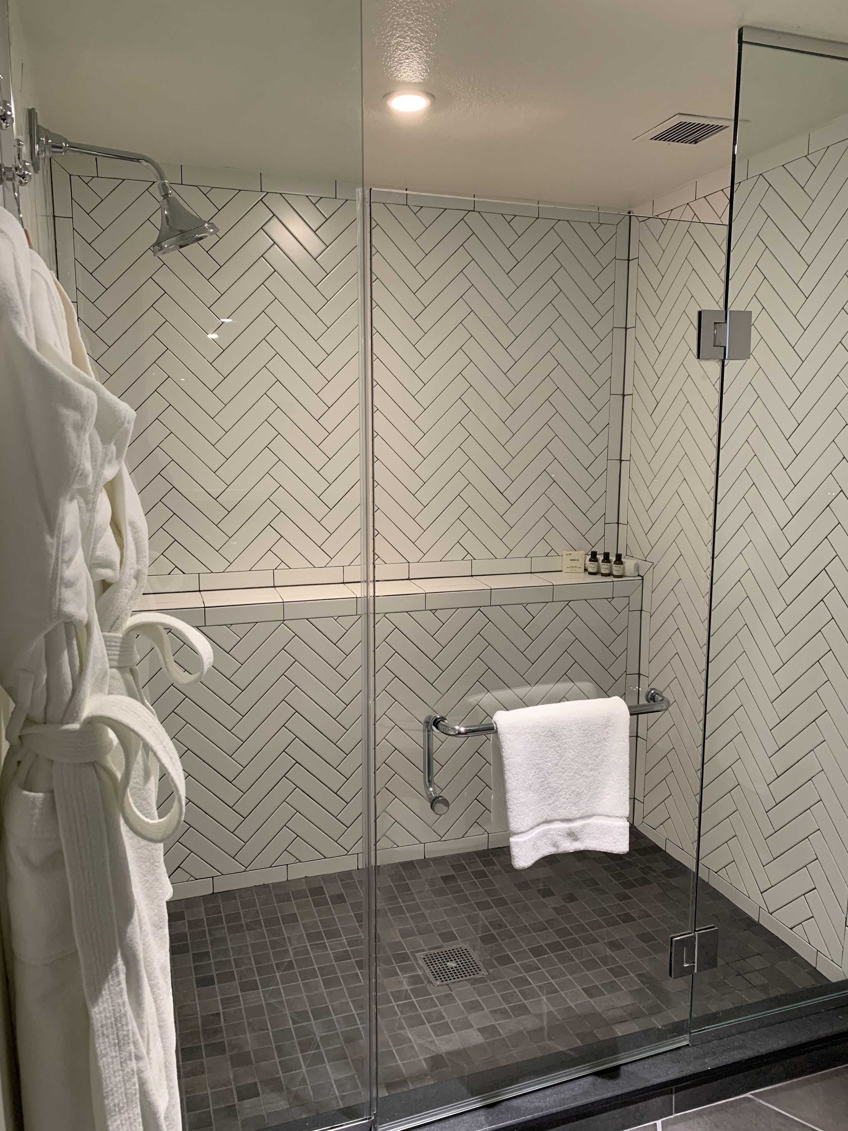 Un espace douche moderne avec une porte en verre, une serviette blanche accrochée au mur et un pare-splash en carreaux disposés en motif chevrons.