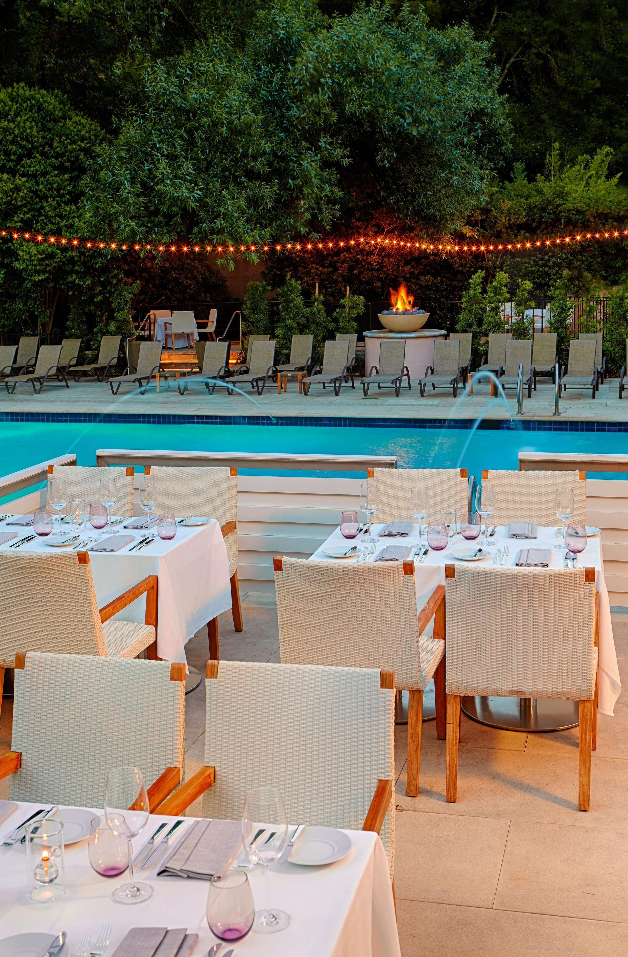 Un espace de restauration aménagé au bord de la piscine, avec des tables pour deux, entouré de chaises longues et illuminé par une guirlande lumineuse.