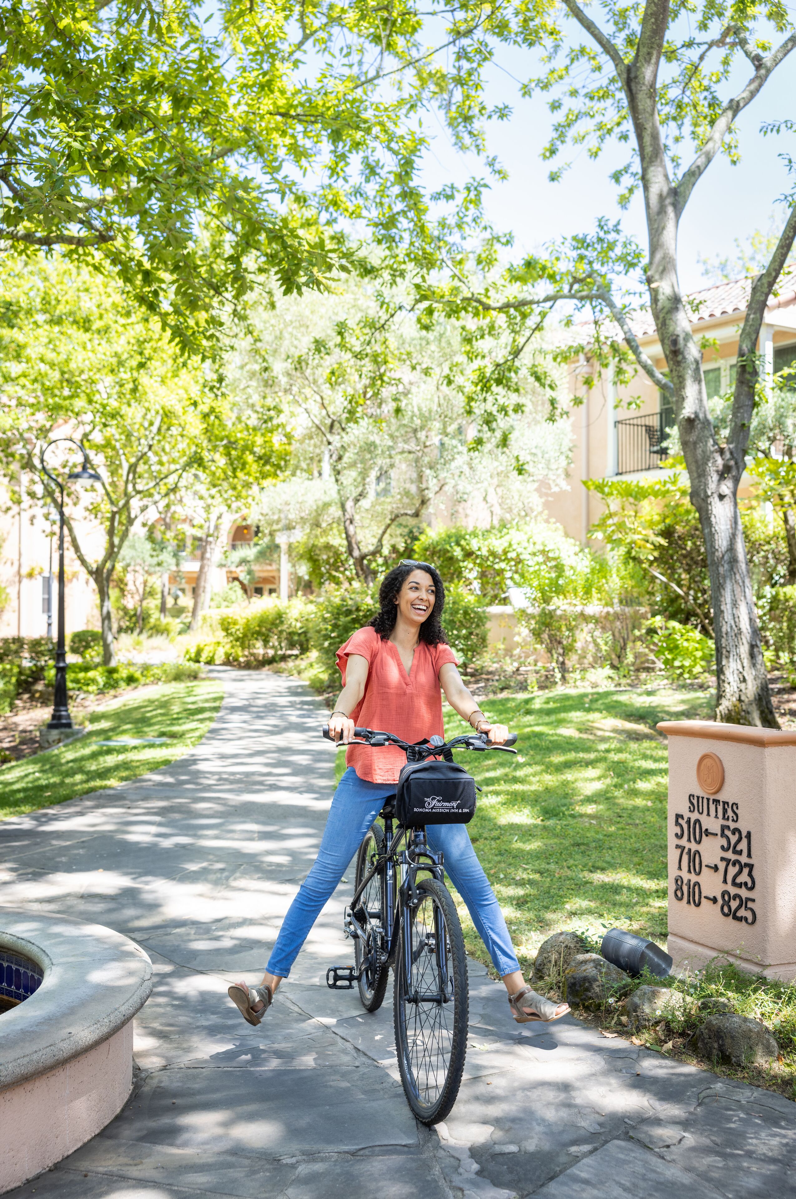 A woman riding a bicycle on a paved path in a well-maintained garden area with trees and a building in the background.
