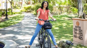 A woman riding a bicycle on a paved path in a well-maintained garden area with trees and a building in the background.