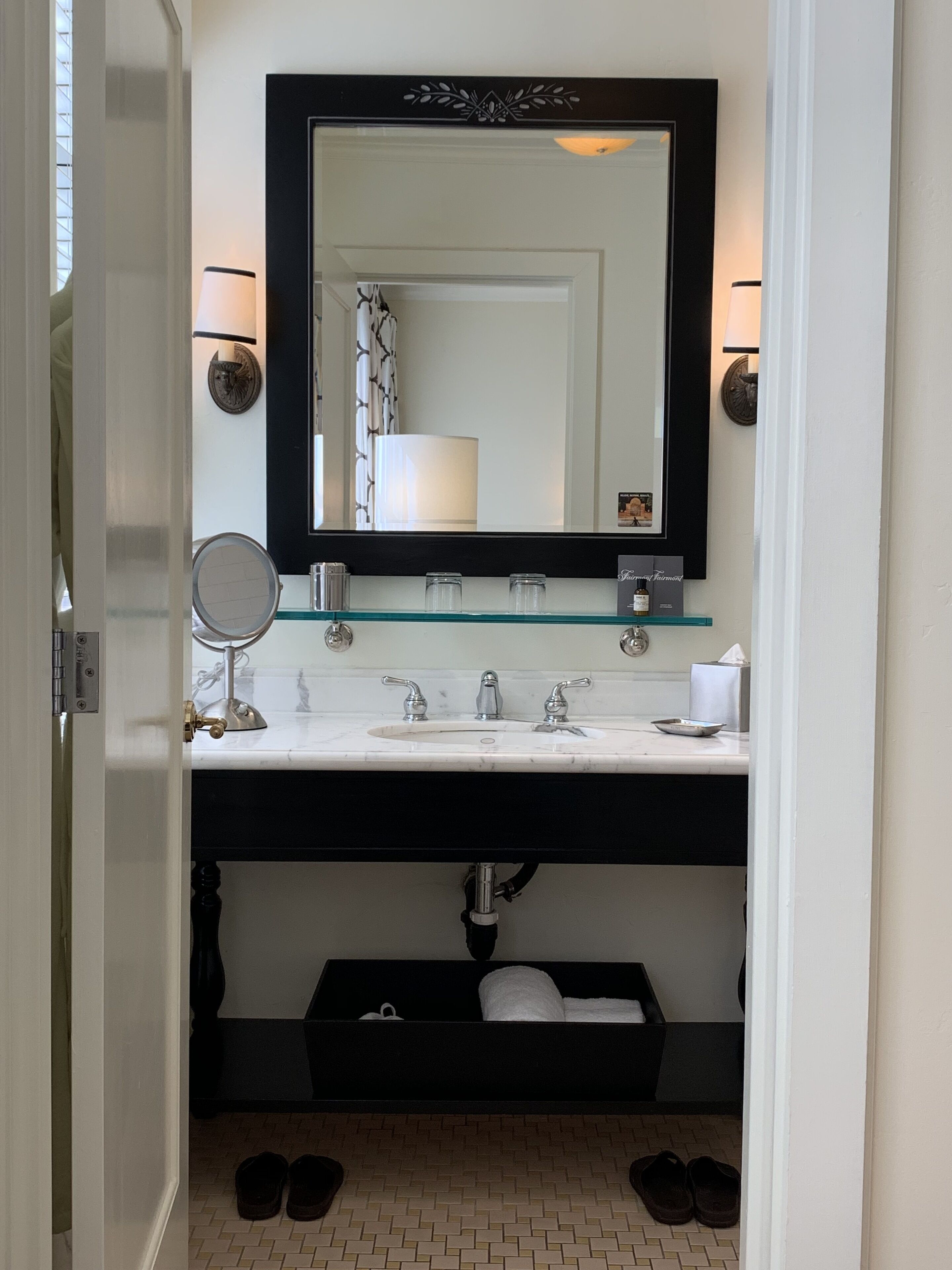 A bathroom with a white sink, black vanity, and a large mirror. There is a towel rack, a small round mirror, and a tissue box on the vanity.