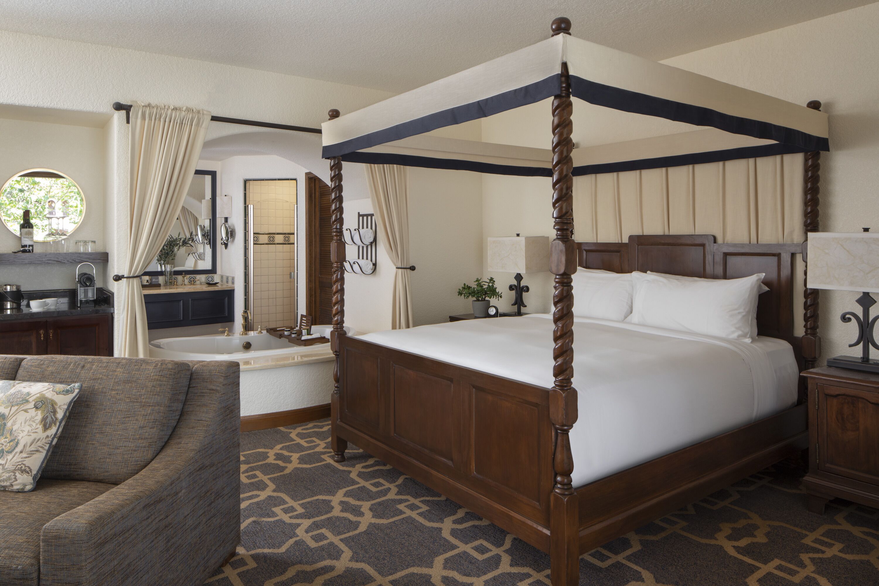 A four-poster bed with a canopy, a sitting area with a chair, a bathroom with a bathtub, and a mirror.
