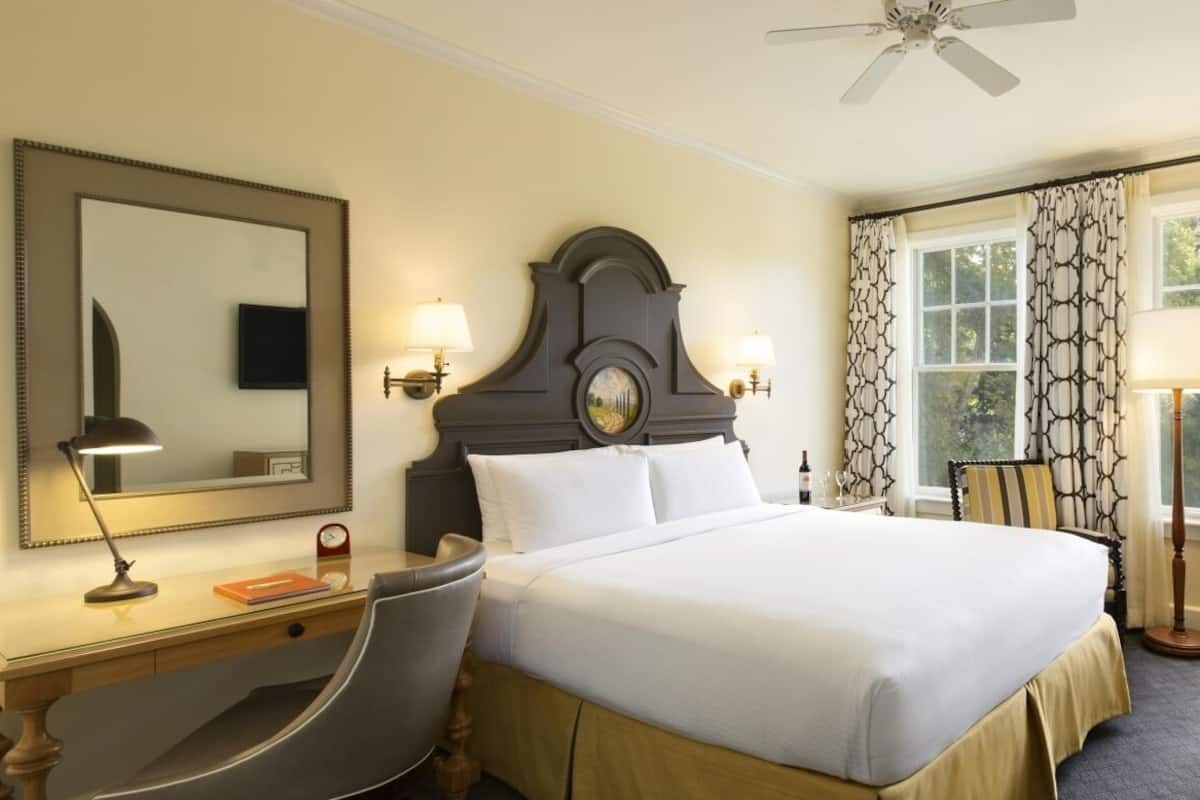 Una habitación de hotel con una cama grande, un escritorio con una silla, una lámpara de escritorio, un televisor montado en la pared y una ventana con cortinas estampadas.