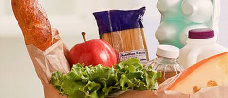 A paper bag containing a baguette, an apple, lettuce, a package of pasta, a bottle of milk, and a carton of eggs.