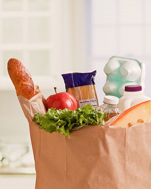 Una bolsa de papel con una baguette, una manzana, lechuga, un paquete de pasta, una botella de leche y una cartona de huevos.