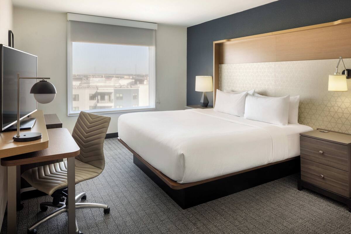 A hotel room with a bed, desk, chair, and a window with a city view.