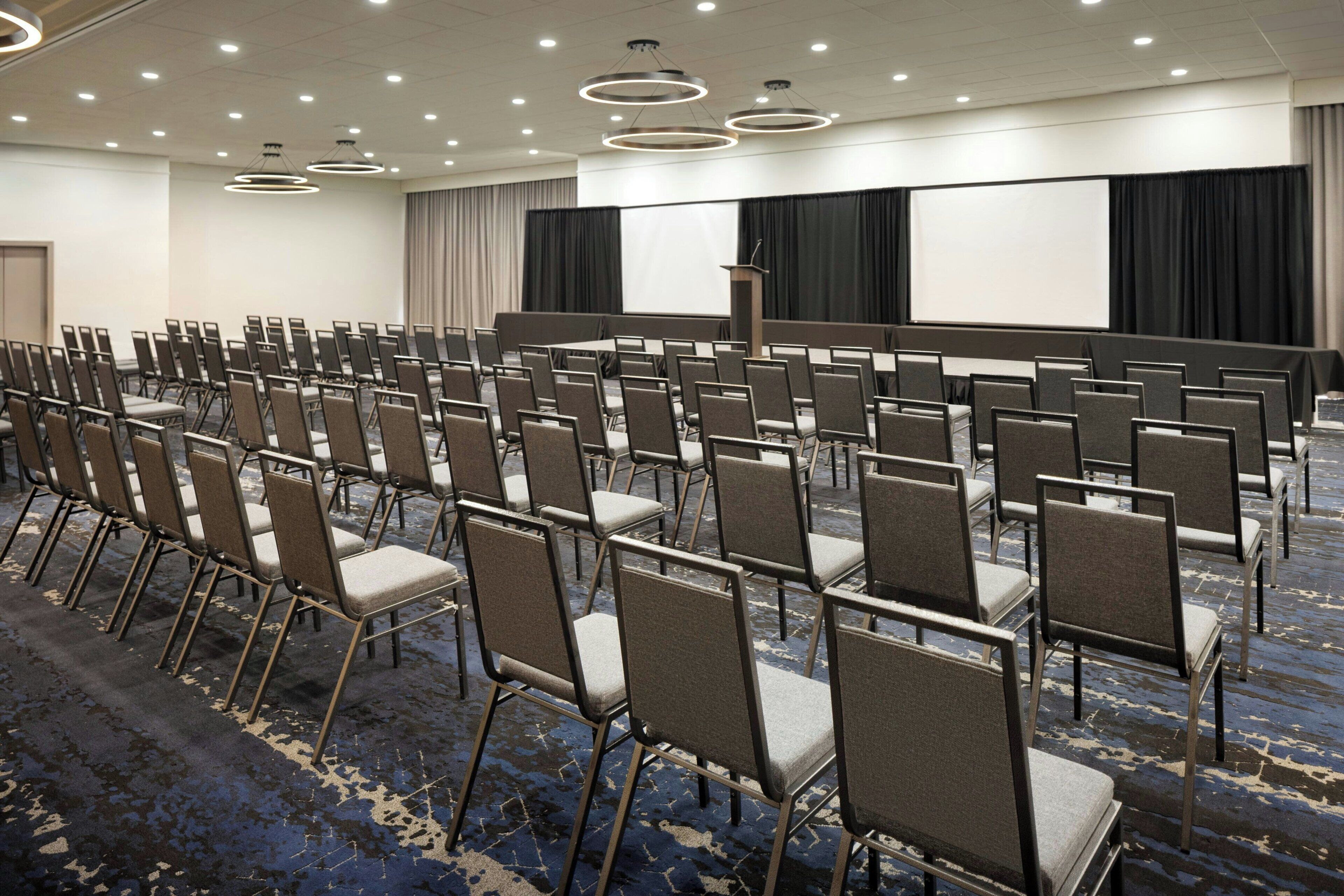 A conference room with rows of chairs, a podium, a large screen, and a carpeted floor.