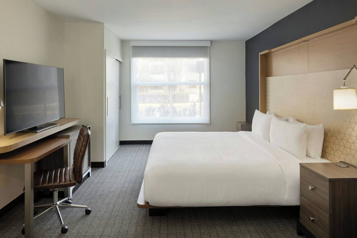 A hotel room with a bed, a desk with a TV, a chair, a nightstand, and a window.