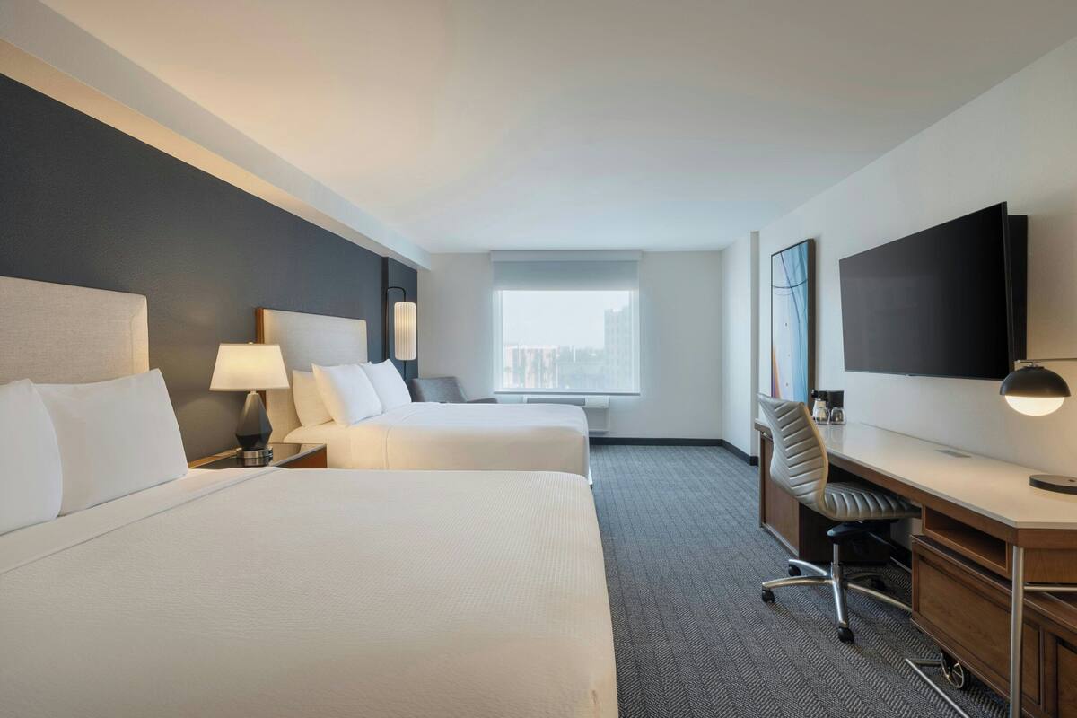 A hotel room with two beds, a desk, a chair, a TV, and a window with a city view.
