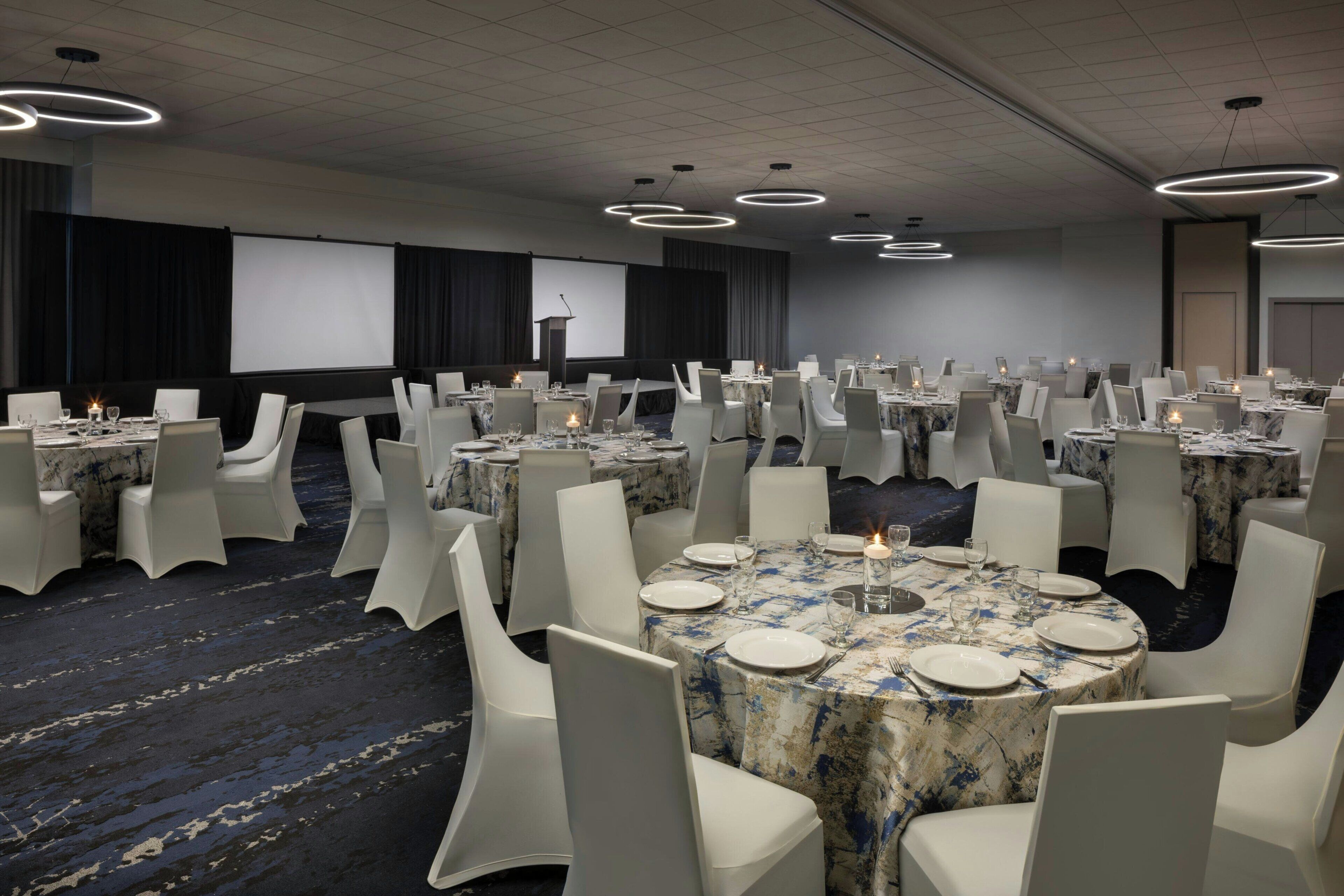 Une salle de conférence avec des tables rondes, des chaises blanches et un écran de projection.
