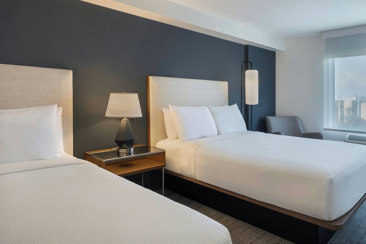 A hotel room with two beds, a nightstand with a lamp, a chair, and a large window offering a city view.