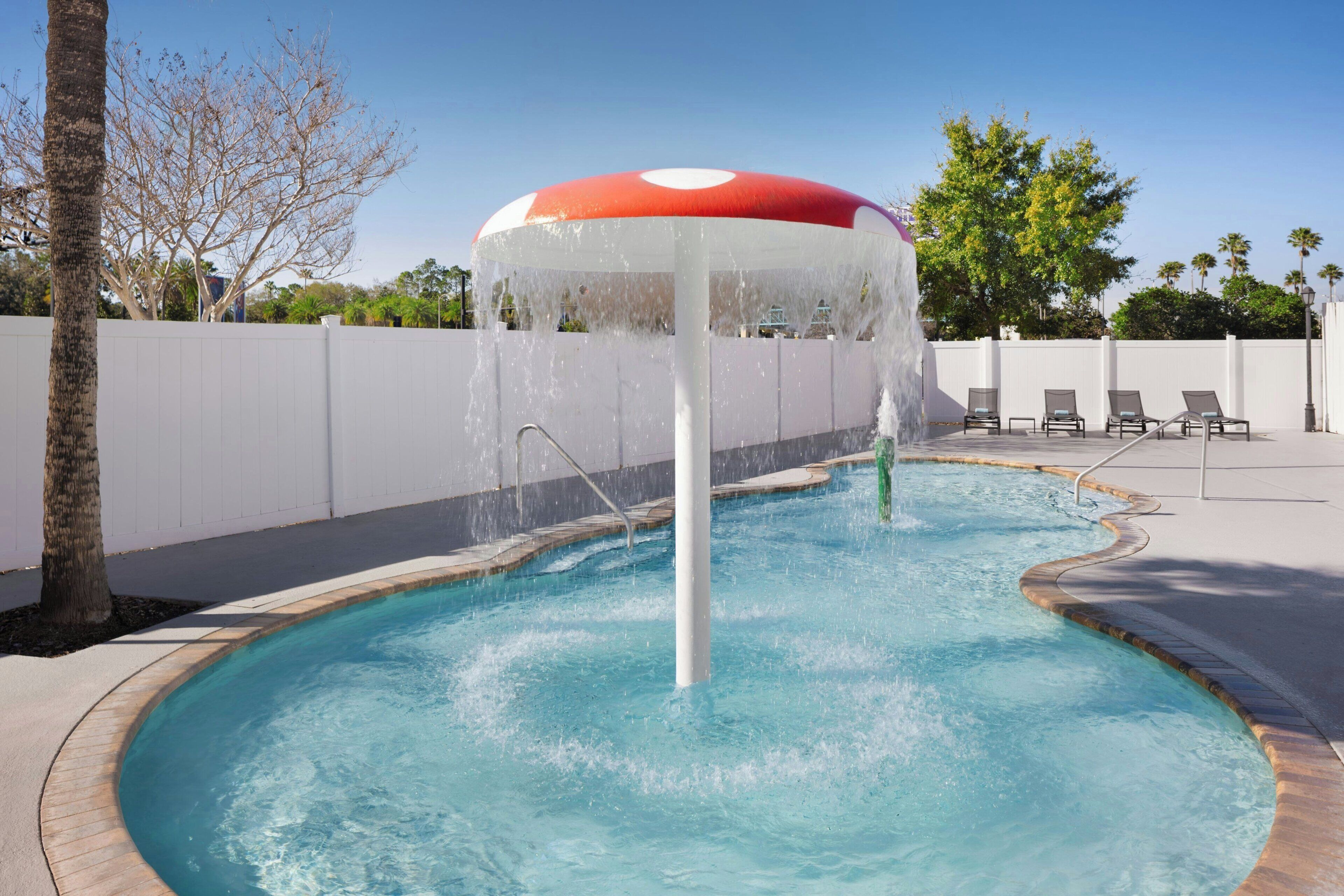 Ein Swimmingpool mit einer Springbrunnenanlage, umgeben von einer weißen Mauer und Palmen.